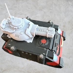 Peut inclure: Une tourelle de char imprimée en 3D de couleur grise avec un long canon et une antenne relevée. La tourelle est montée sur un châssis de char noir et rouge.