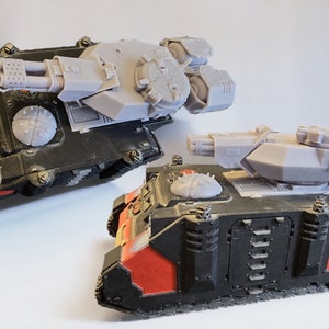 Puede incluir: Dos vehículos de combate blindados en miniatura con cascos pintados en negro y rojo y torretas de plástico gris claro sin pintar. Cada tanque cuenta con un sistema de armas de varios cañones, una escalera lateral y una escotilla.