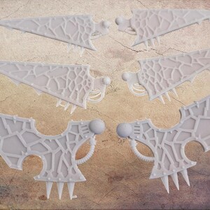 May include: Six light grey, wing-shaped objects with a textured, web-like pattern. Each wing has a rounded sphere and sharp, pointed protrusions. The objects are arranged on a textured, brown surface. The objects appear to be 3D printed.