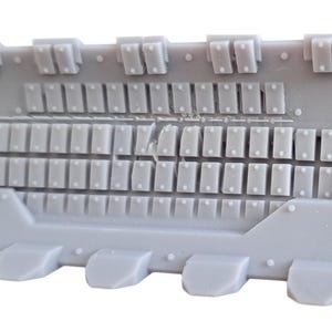 Op de afbeelding: Een grijs plastic model van een ruimteschip met een gedetailleerd exterieur met een raster van kleine vierkanten en een rij kleine, rechthoekige uitsteeksels langs de onderkant.