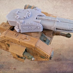 May include: A detailed model of a tank, featuring a light gray turret and a tan body. The turret has a large cannon and is adorned with a winged skull emblem. The tank's body has visible panel lines and hatches.
