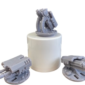 May include: Three gray miniature turrets, likely for tabletop gaming or model building, are displayed against a white background. One turret is mounted on a white cylinder. The turrets feature detailed designs with multiple barrels and mechanical elements.