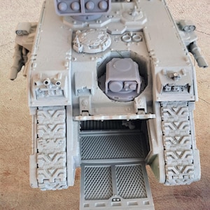 Puede incluir: Maqueta de tanque gris con orugas detalladas, lanzamisiles y una escotilla abierta. El vehículo en miniatura tiene una superficie texturizada y detalles intrincados, lo que sugiere un tema militar o de ciencia ficción. La escotilla revela el interior.