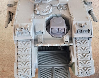 Może przedstawiać: Szary model czołgu z szczegółowymi gąsienicami, wyrzutniami rakiet i otwartym włazem. Miniaturowy pojazd ma teksturowaną powierzchnię i skomplikowane detale, sugerujące motyw militarny lub science fiction. Właz odsłania wnętrze.