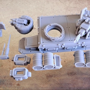 Può includere: Una collezione di parti di modelli in plastica grigio chiaro, tra cui una grande base rettangolare con un'apertura circolare, due torrette di cannone, barili e altri componenti. Si tratta di parti di modelli in miniatura.