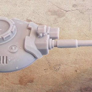 May include: A light gray miniature tank turret with a long barrel. The turret features detailed mechanical components, including a circular hatch, vents, and various protrusions. The overall design suggests a model for tabletop gaming or hobby projects.