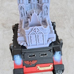 May include: A 3D printed model of a gothic cathedral with a detailed design, featuring a pointed archway and a series of small towers. The model is painted in a light grey colour and is mounted on a black and red base.