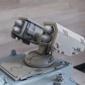 May include: A light grey plastic model of a military vehicle turret featuring two rectangular missile launcher pods mounted on a rotating base. Each pod contains two visible missile tips, and the structure includes moulded panel details.