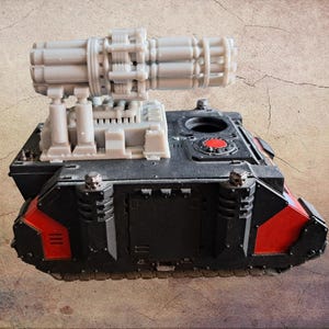 Pode incluir: Um modelo de tanque preto e vermelho com uma grande arma cinza de vários canos montada no topo. O tanque tem detalhes em vermelho e um detalhe circular vermelho na parte superior. O modelo está sobre um fundo texturizado e bege.