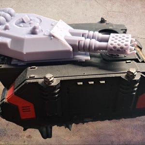 Puede incluir: Maqueta de tanque en miniatura, principalmente negra con detalles rojos, y una torreta gris claro. La torreta presenta dos cañones grandes y una sección superior detallada. El modelo se encuentra sobre un fondo texturizado en tonos sepia.