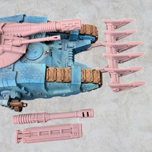 May include: A pink and blue plastic model of a tank with a large claw on the side. The tank has a large cannon on top and a smaller cannon on the front. There are two additional pink plastic pieces, one long and one short, that appear to be weapons.