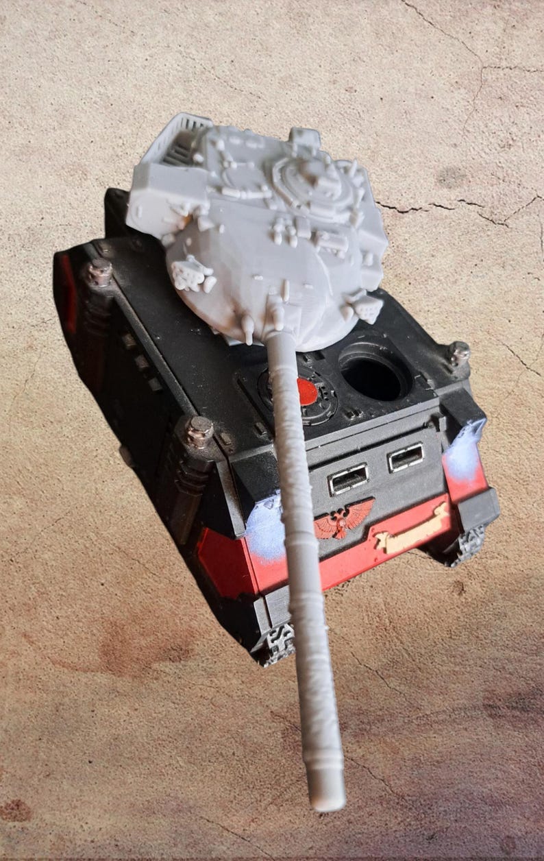 May include: A miniature tank model featuring a black and red chassis with a prominent red eagle emblem on the front. The tank has a long, light gray turret barrel and a light gray turret top, with a small circular red detail on the upper deck.