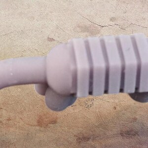 May include: A light purple, stylized miniature car exhaust system. The exhaust pipe curves to the left, connected to a rectangular muffler with vertical ridges. The object is a solid, single-piece design.