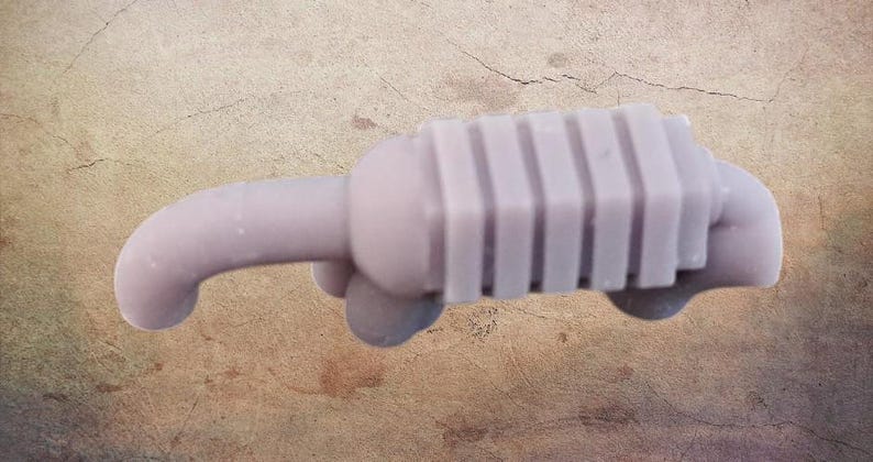 May include: A light purple, stylized miniature car exhaust system. The exhaust pipe curves to the left, connected to a rectangular muffler with vertical ridges. The object is a solid, single-piece design.