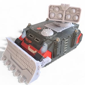 Pode incluir: Um tanque em miniatura preto e vermelho com uma lâmina branca e um lançador de mísseis cinza no topo. O tanque tem uma faixa vermelha e um símbolo branco no lado.