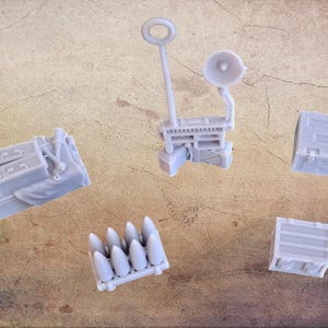 May include: A selection of small, pale grey miniature military models. The set includes a radio, ammunition, crates, and other equipment. The items are arranged on a textured, brown surface.