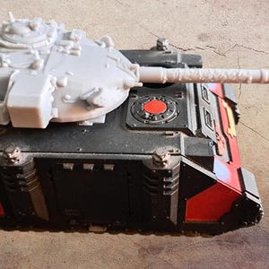 May include: A miniature tank model featuring a black and red chassis with a light gray turret and a long barrel. The turret includes a small white railing on the side, while the main body displays a circular red emblem on the top surface.