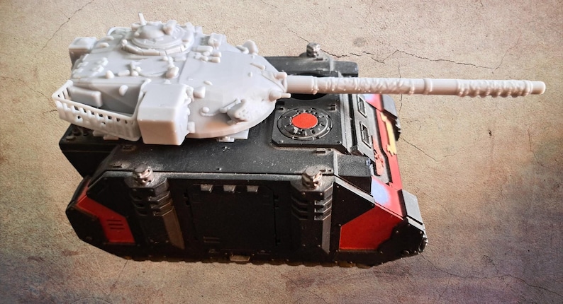 May include: A miniature tank model featuring a black and red chassis with a light gray turret and a long barrel. The turret includes a small white railing on the side, while the main body displays a circular red emblem on the top surface.