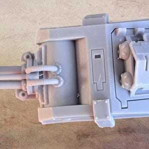May include: A grey plastic model of a futuristic weapon with a large barrel and a smaller, boxy turret on top. The weapon has a detailed design with pipes and vents.