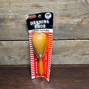 May include: A vintage darning knob made of selected hardwood, still in its original packaging. The packaging is black and red with the text "Darning Knob" and "For Socks, For Gloves". The knob is a light brown color.