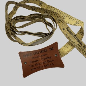 May include: An antique measuring tape with black markings and the text "FAMOUS-STERNBERG, INC." on the tape and a brown leather tag. The tag reads "Summer Clothing For Men and Boys NEW ORLEANS, LA."