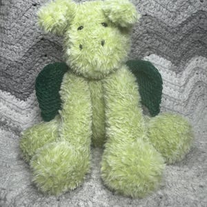 May include: A plush, lime green dragon stuffed animal with dark green wings. The dragon has a fluffy texture and is sitting on a gray and white crocheted blanket. The dragon's features include small black eyes and a stitched nose.