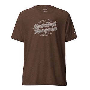 May include: A brown t-shirt with the text "Skyline Trail Switchback Renegades CORONA, CA" in a white, stylized font. The shirt has a crew neck and short sleeves. The fabric appears to be a soft, heathered material.