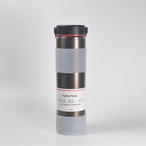 Puede incluir: Una botella de agua de acero inoxidable con tapa negra y funda de silicona gris. La botella tiene un acento rojo en la tapa y el texto "Hydro Flask" y "Trail Series" impreso en el lateral. La botella es de 750 ml (24 oz).