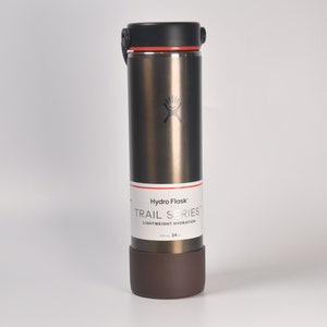 Puede incluir: Una botella de agua Hydro Flask Trail Series de acero inoxidable marrón con tapa negra y base de silicona marrón. La botella tiene una capacidad de 709 ml (24 oz).