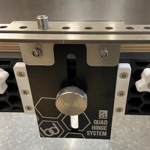 May include: A black and silver woodworking jig with white knobs and the text "QUAD HINGE SYSTEM".