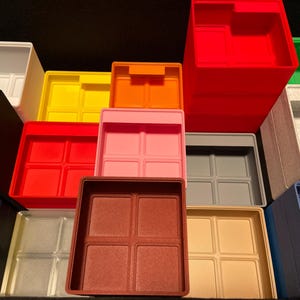 May include: A collection of colorful plastic storage bins in various shades, including red, pink, yellow, and blue. Each bin has a square shape with a divided interior, suitable for organizing small items. The bins are stacked and arranged, showcasing their different colors and sizes.