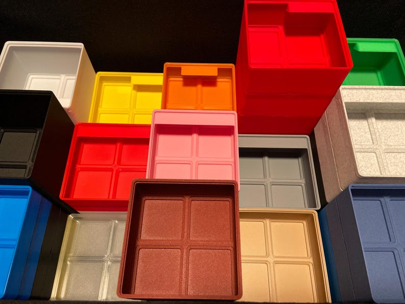 May include: A collection of colorful plastic storage bins in various shades, including red, pink, yellow, and blue. Each bin has a square shape with a divided interior, suitable for organizing small items. The bins are stacked and arranged, showcasing their different colors and sizes.
