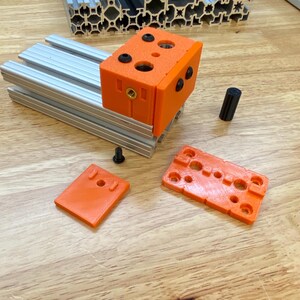 May include: Orange 3D printed parts for a construction project. The parts include a block with holes, a small rectangle, and a larger rectangle with multiple holes.
