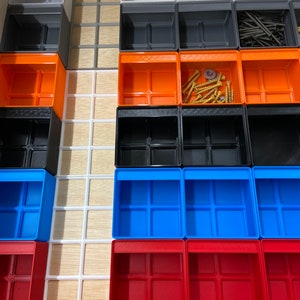 May include: A drawer organizer system featuring a variety of colorful, small, open-top storage bins. The bins are in shades of gray, orange, black, blue, and red. Some bins contain screws and other small hardware. The organizer is set in a wooden drawer.