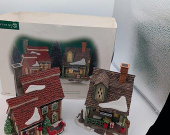 May include: Two miniature ceramic houses with snow-dusted roofs and chimneys, featuring red and brown shingled textures, green window frames, and festive accents. One house includes a red sleigh, and the other is labeled "Walden Cottage" on its porch.