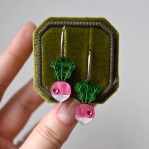 Op de afbeelding: Zilveren oorringen met een radijsontwerp. Elke oorring heeft een roze en witte radijs met groene bladeren en een kleine rode edelsteen. De oorbellen worden gepresenteerd in een groene fluwelen sieradendoos.