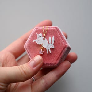 Könnte beinhalten: Eine zarte goldene Kettenkette mit einem weißen Porzellan-Hasenanhänger und einem rosa Kristall-Sternanhänger. Die Kette wird in einer rosa Samtbox präsentiert.