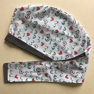 May include: A white surgical scrub cap with a repeating pattern of black line-drawn breasts and red hearts. The cap has a dark gray band and ties. The fabric is a light, neutral color.