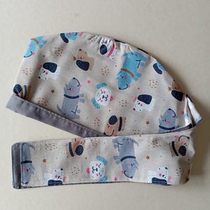 May include: A beige and gray scrub hat with a tie. The hat has a pattern of cartoon dogs in various colors and poses. The tie is made of the same fabric as the hat.