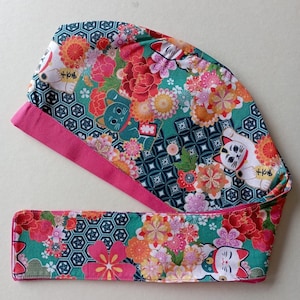 May include: A pink and teal fabric headband with a floral pattern and images of lucky cats. The headband has a matching fabric tie that can be tied around the head.