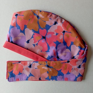 Puede incluir: Un gorro quirúrgico con estampado floral y corbata a juego. El gorro presenta un diseño vibrante de flores rosas, naranjas y moradas sobre un fondo azul. La corbata y el ribete son de color coral liso. Diseñado para profesionales de la medicina.