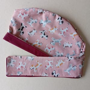 May include: A pink surgical scrub hat with a pattern of colorful dogs. The hat has a maroon trim and a tie.