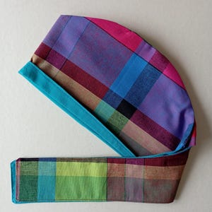 May include: A colorful plaid fabric headband with a turquoise trim. The headband is tied in a knot at the back of the head.