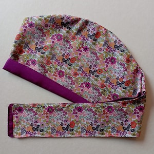 May include: A purple and white floral print scrub hat with a purple satin trim. The hat has a tie that is also made of the same floral print fabric.