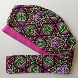 May include: A patterned scrub cap with a matching headband. The cap and headband feature a geometric design in green, pink, and black. The trim is a solid magenta color. The cap is designed to be worn in a medical or professional setting.