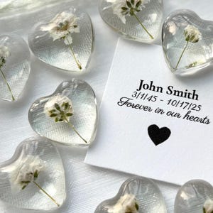 Peut inclure: Des presse-papiers en forme de cœur transparents avec des fleurs blanches. Une carte indique "John Smith 3/1/45 - 10/17/25 Forever in our hearts" avec un cœur noir. Les presse-papiers sont sur un fond blanc.