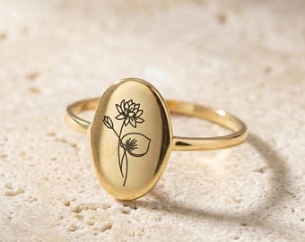Puede incluir: Un anillo dorado con una cara ovalada que presenta un diseño floral negro. El anillo tiene una banda delgada y un acabado pulido. El diseño floral incluye una flor y hojas. El anillo está sobre un fondo neutro.