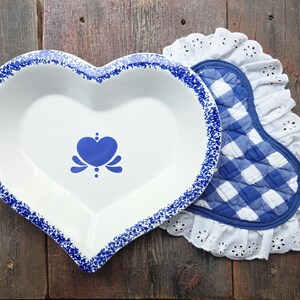 May include: A heart-shaped white ceramic dish with a blue speckled rim and a central blue heart icon, paired with a heart-shaped quilted potholder featuring a blue and white gingham pattern and a white eyelet lace ruffled trim border.