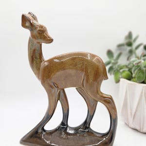 May include: A standing deer figurine crafted from ceramic with a glossy finish. The deer features a gradient color scheme, transitioning from a light brown on the upper body to a deep, dark brown at the base of the legs and the bottom edge.