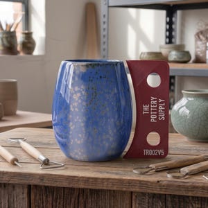 Può includere: Una tazza in ceramica blu con finitura maculata e un manico piatto color bordeaux con due fori circolari e la scritta THE POTTERY SUPPLY TR002V5. La tazza è posizionata su una superficie in legno insieme a vari strumenti per la ceramica.